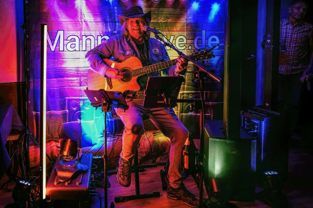 Mannie Live sitzend mit Gitarre vor Mannie-Live-Banner und Bühnenequipment